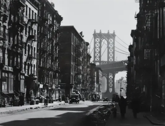 Black and white photograph of city buildings with the Brooklyn bridge in the background between the rows of buildings.