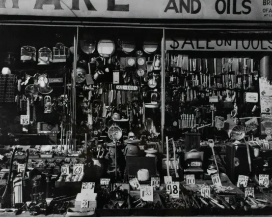 Black and white photograph of a clutters hardware store storefront with price tags, windows, and a sale sign in the window.