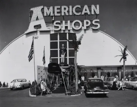 Photograph of a neon letter sign that reads "American Shops" in the parking lot with large shopping center behind.