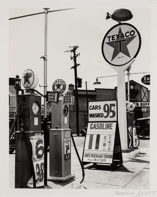 Black and white photograph of a gas station named Texaco with two gas pumps on the left and a sign on the right.