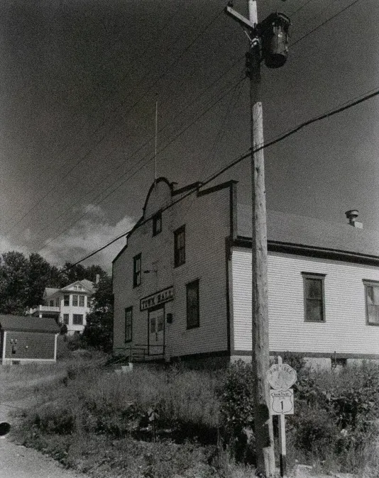 Back and white photograph of a telephone pole and wires in the foreground with a house in the background.
