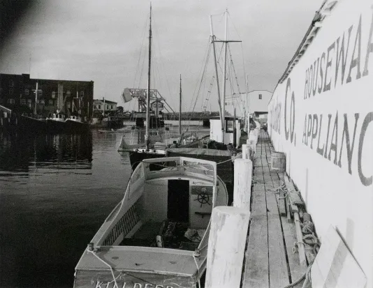 Black and white image of a dock with a boat on the left and a sign on the wall on the right titled "Houseware Appliances"