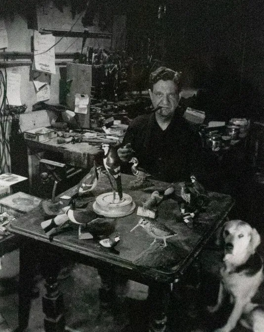 Black and white photo of a man sitting at a work table with a pipe in his mouth and a dog sitting to his left.