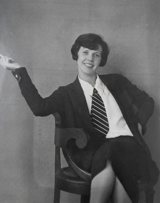 Woman wearing a blazer and tie sitting in a chair with her right hand in the air and her left hand on her hip.