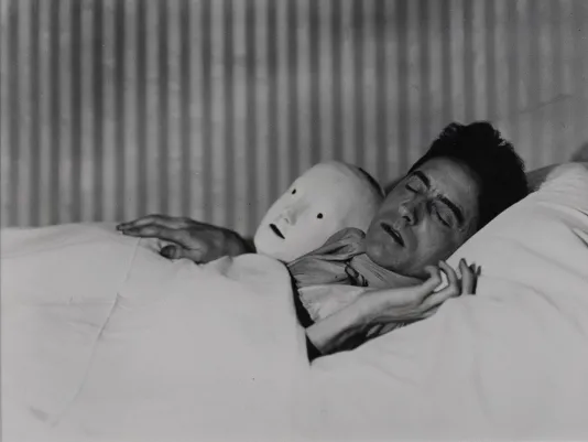 Black and white photograph of a person sleeping in bed with a white mask next to them.