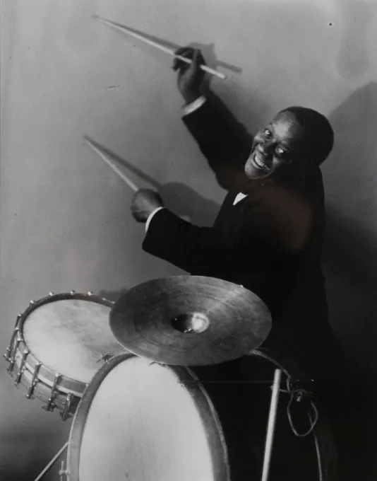 A man sits at a drum set and plays the drums while he smiles and tilts his head to the left.