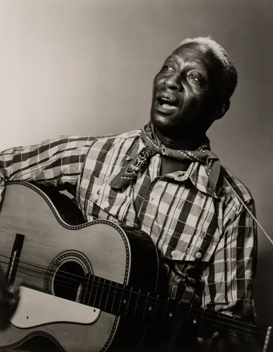 A black man with a plaid shirt playing the guitar and singing.