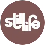 Logo for Still Life Studios with a brown circle and white text. Text reads &quot;Still Life&quot;